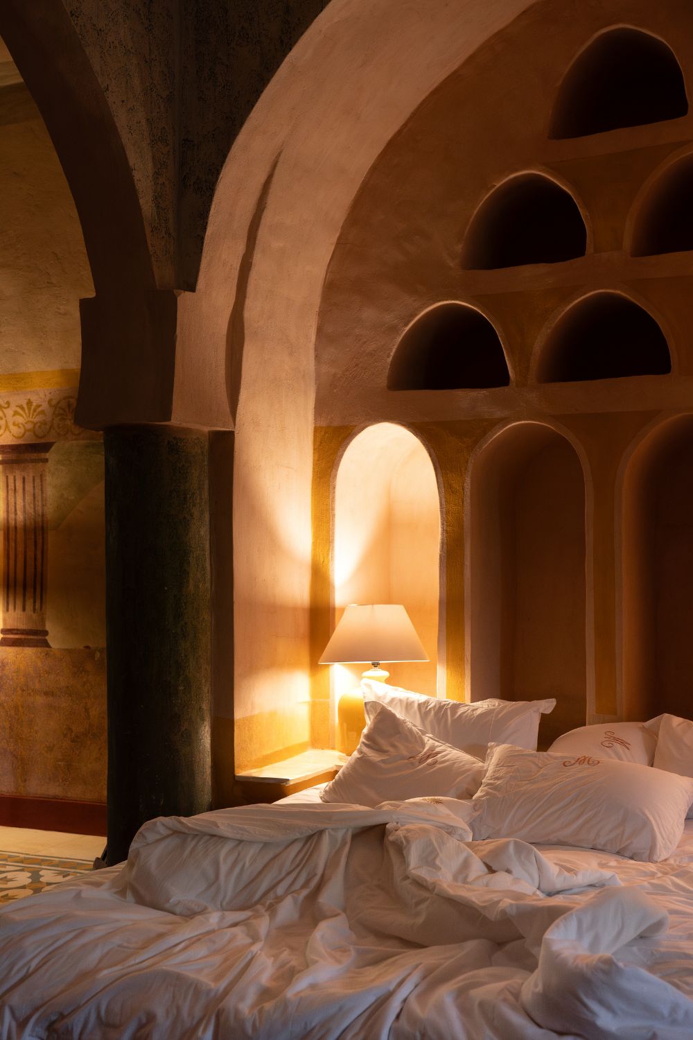 Warm lamplit bedroom with arched plaster niches and rumpled white linens