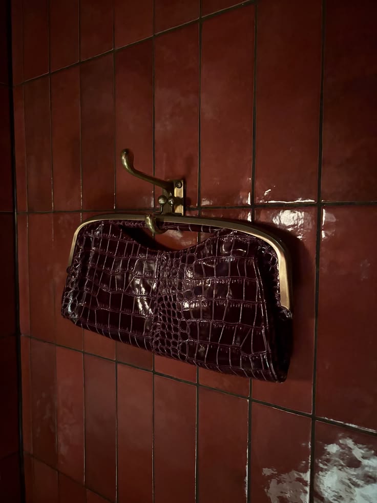 Burgundy crocodile-embossed clutch hanging on a brass hook against red tile