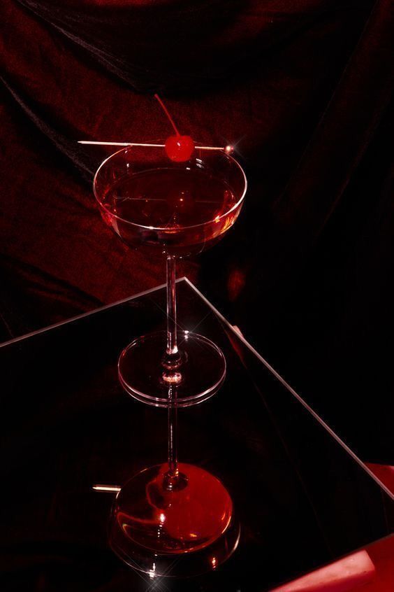 Dark cocktail coupe with a cherry on a mirrored table against red velvet